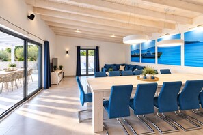 A modern dining area with a large table and blue chairs, a kitchen area with a TV and counter, and a view of the sea through large windows. - Wellness & Spa, Villa STELLA - heated pool, close to beaches, playground, Istria (PULA)