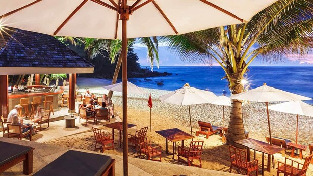 Un restaurante en la playa con terraza, bar y sombrillas que dan sombra.