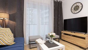 A modern living room with a blue sofa, a white coffee table, a TV on a wooden stand, and a large window with curtains.
