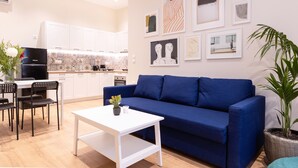 A modern living room with a blue sofa, a white coffee table, and a dining area with a black table and chairs. There is a kitchen in the background with white cabinets and a black refrigerator. - Casa Moderna - Two Bedroom Apartment (Corfu)