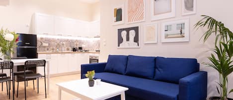 A modern living room with a blue sofa, a white coffee table, and a dining area with a black table and chairs. There is a kitchen in the background with white cabinets and a black refrigerator.