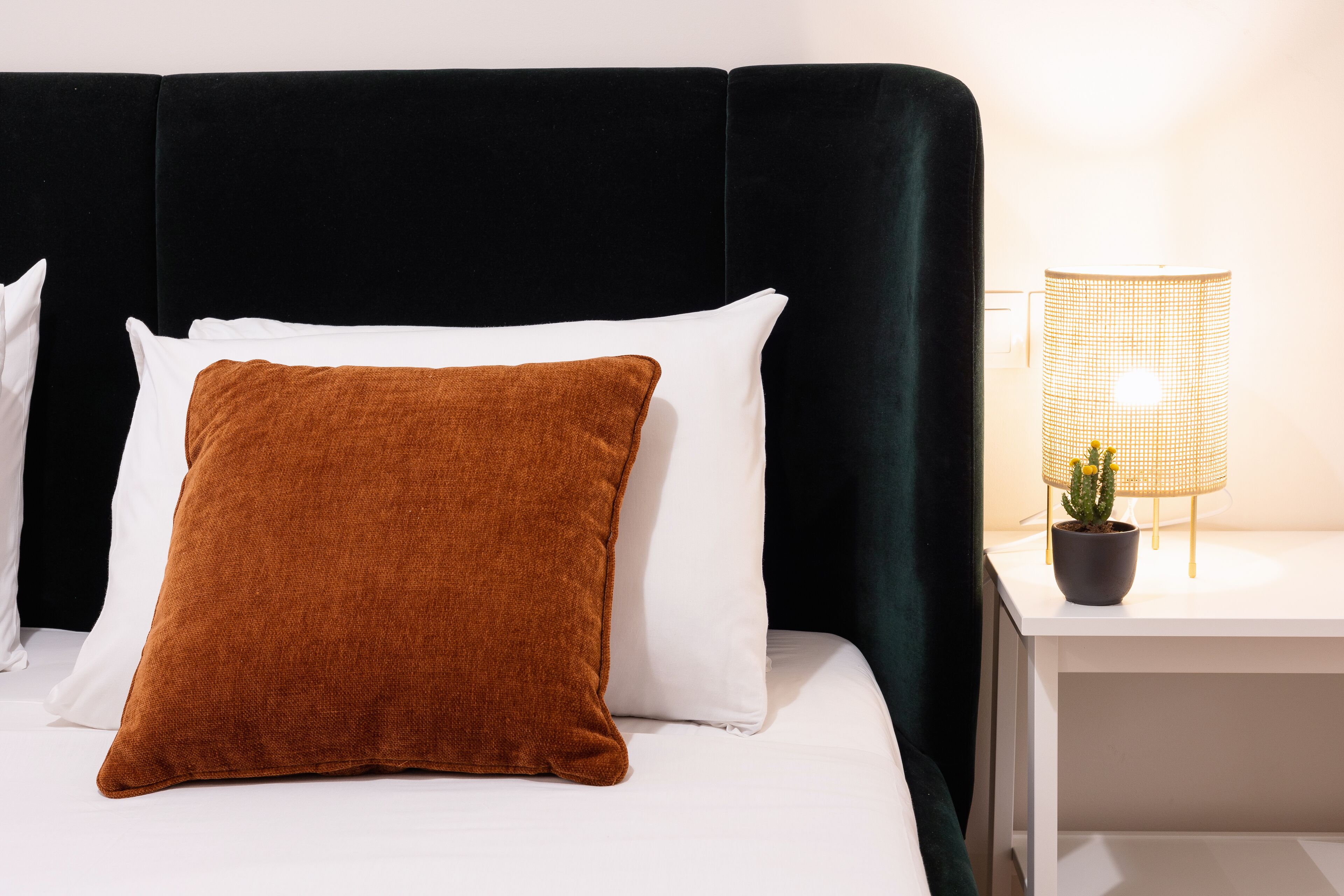 Ein Bett mit schwarzem Kopfteil, weißen und braunen Kissen sowie ein kleiner Tisch mit Lampe und Topfpflanze.