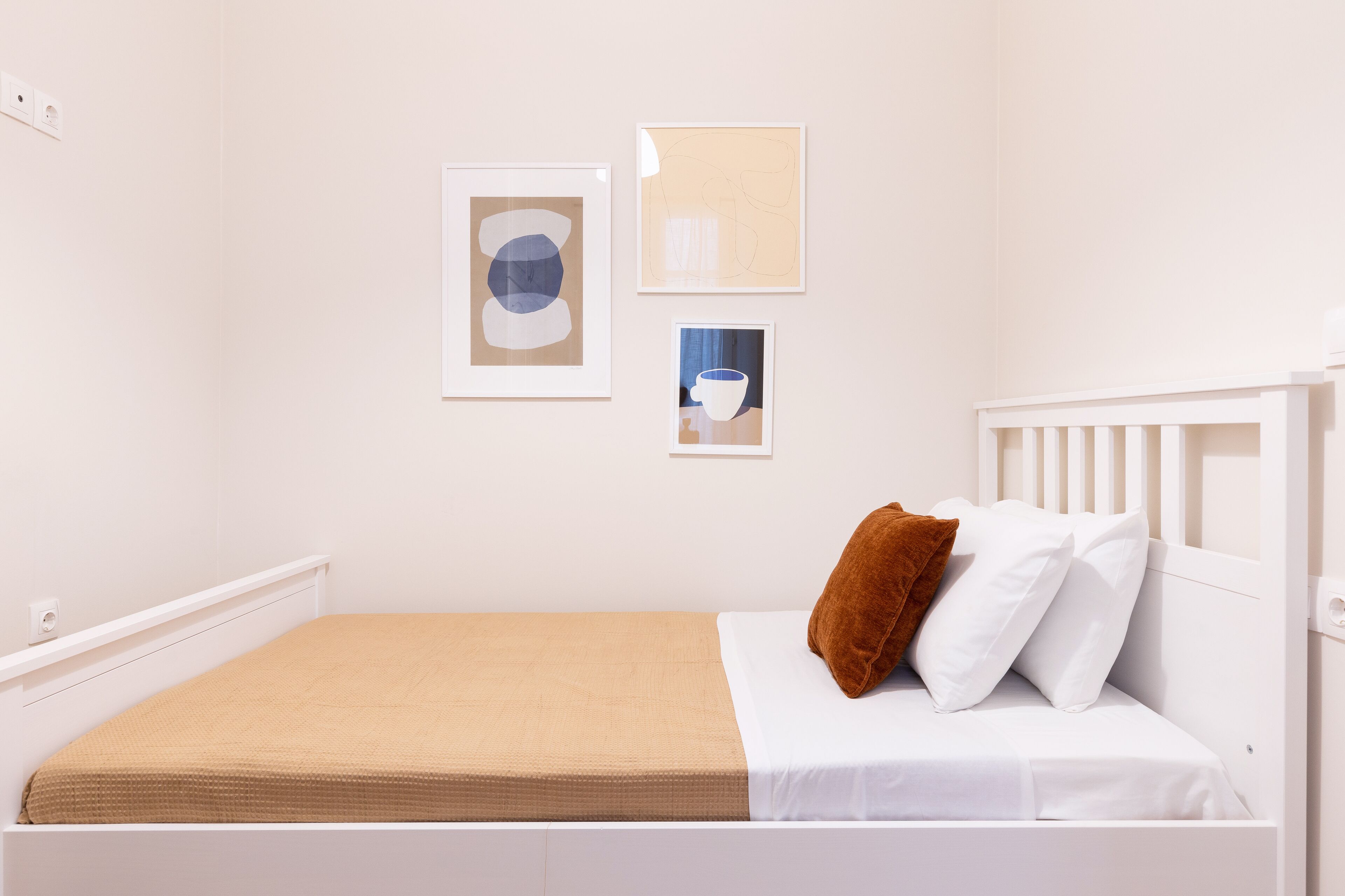 A white bed with a brown blanket, white pillows, and a brown pillow. There are three framed pictures on the wall.