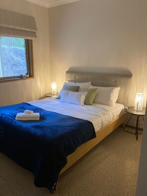 Ein ordentlich bezogenes Bett mit blauer Decke und weißen Kissen, ein Nachttisch mit Lampe und ein Fenster mit Blick auf Bäume.