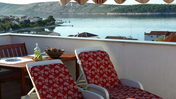 A balcony with two white plastic chairs covered with red floral cushions, a wooden table with a bowl of fruit and a bottle, and a view of a lake with boats and buildings in the background.