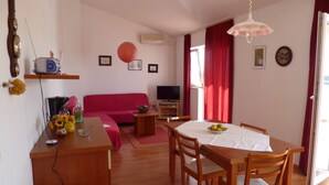 A living room with a red sofa, a TV, a dining table with chairs, and a small kitchen area with a microwave and a kettle.