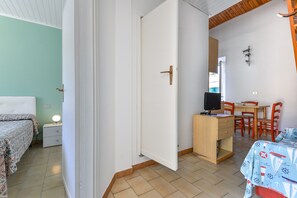 Huoneessa on sänky, työpöytä, tuoli, televisio ja pöytä, jonka päällä on kuvioitu pöytäliina.