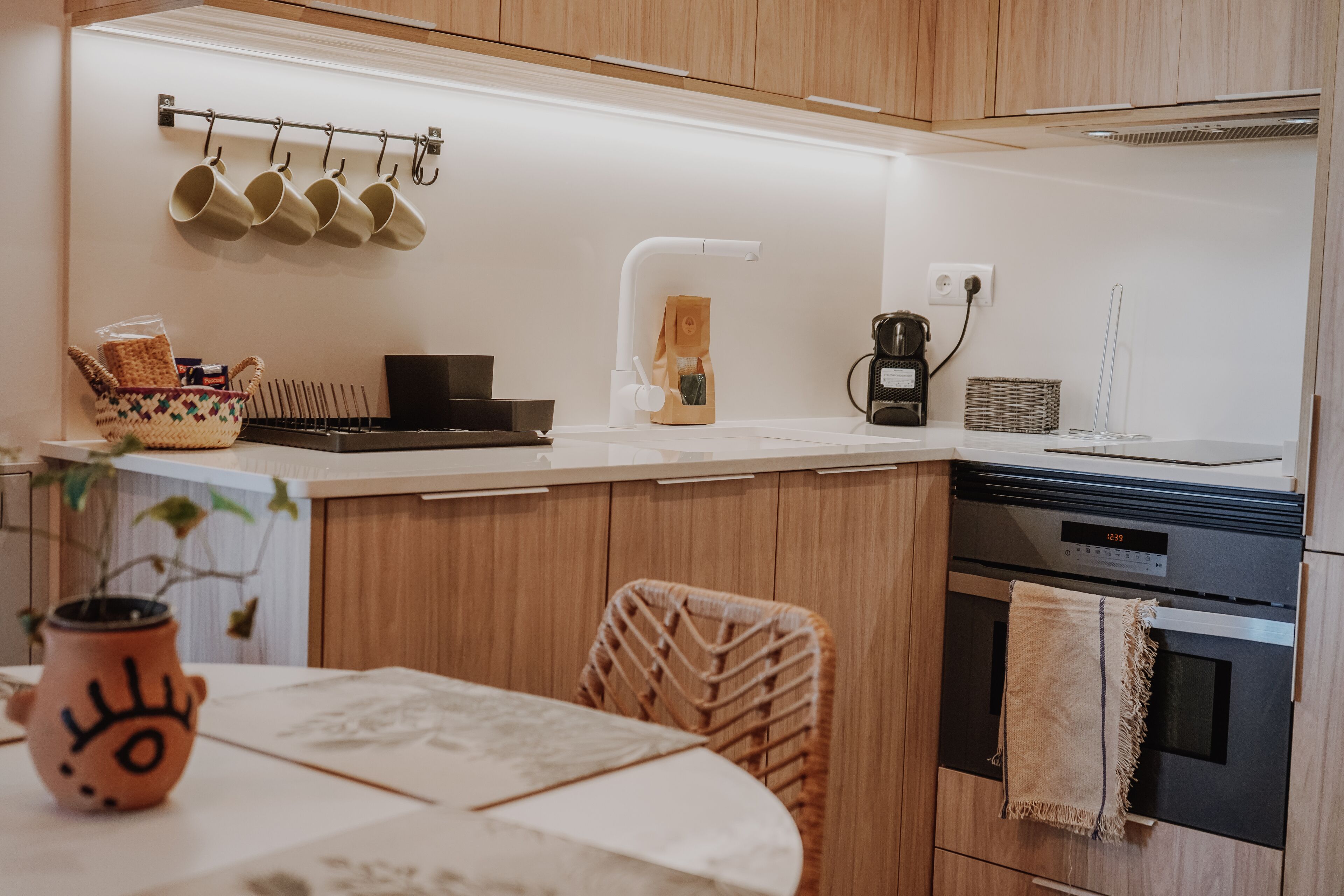 Una cocina moderna con armarios de madera, encimera blanca, un colgador para tazas, una cafetera y un reloj que marca las 12:39.