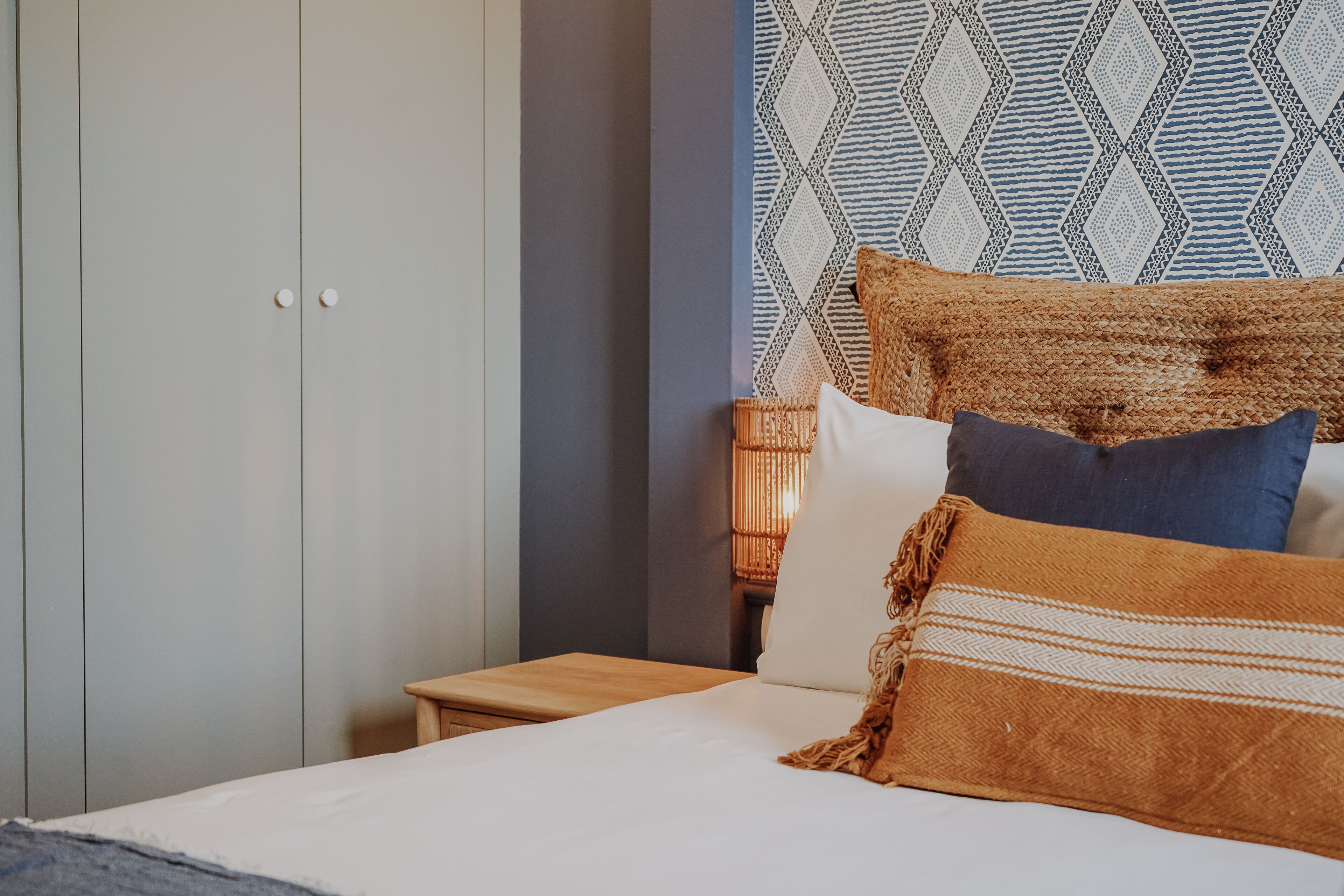 Una cama bien hecha con una manta marrón y almohadas, una mesita de noche de madera y un papel tapiz con un estampado geométrico.