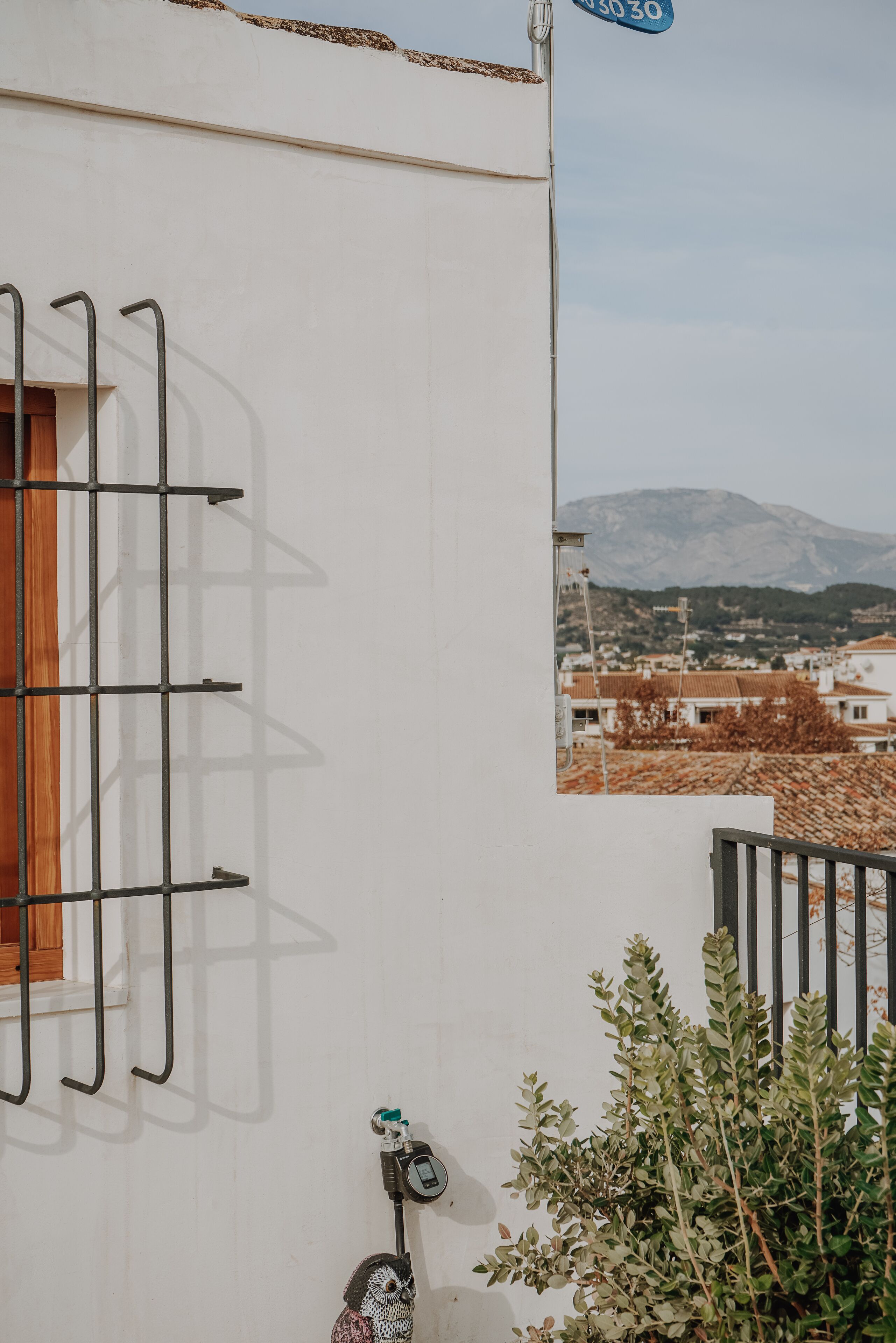 A white building with a metal rack on the wall, a window with a wooden frame, and a view of a mountain and town in the background.