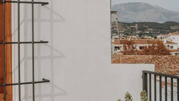 A white building with a metal rack on the wall, a window with a wooden frame, and a view of a mountain and town in the background.