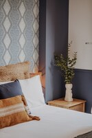 A neatly made bed with multiple pillows, a bedside plant, and a patterned wall behind.