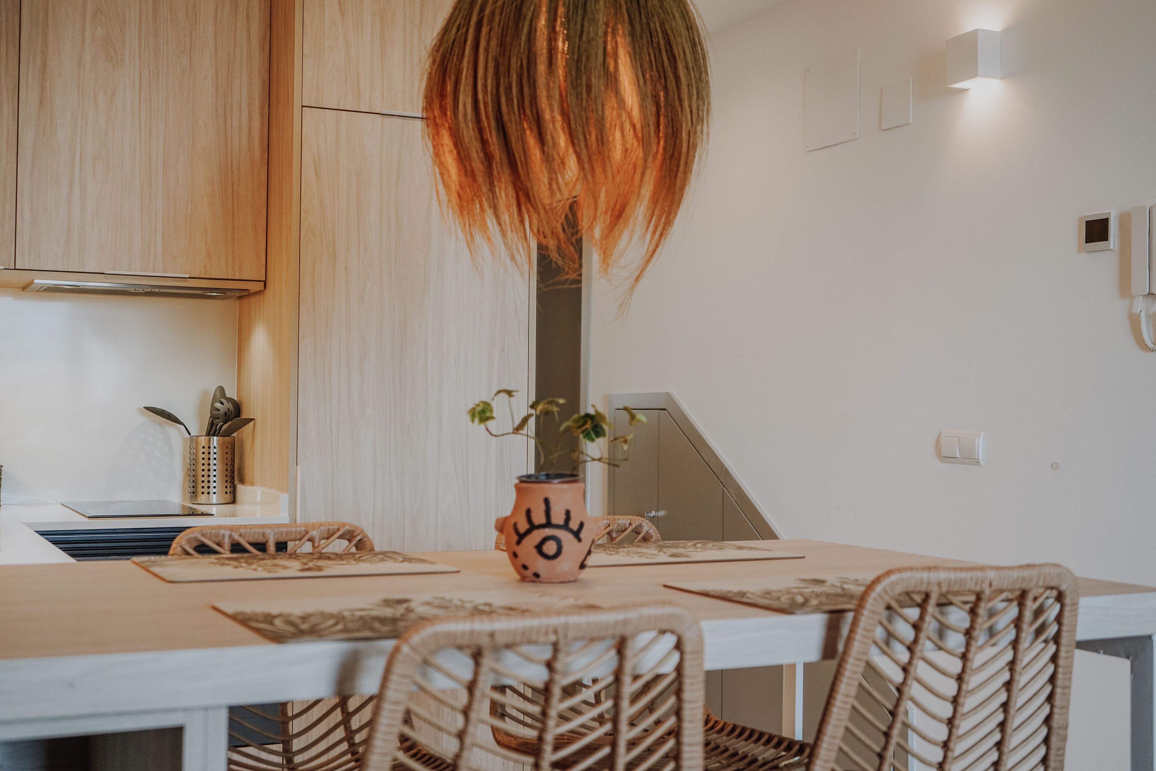 A modern kitchen with wooden cabinets, a laptop, a vase with a plant, and wicker chairs around a table.