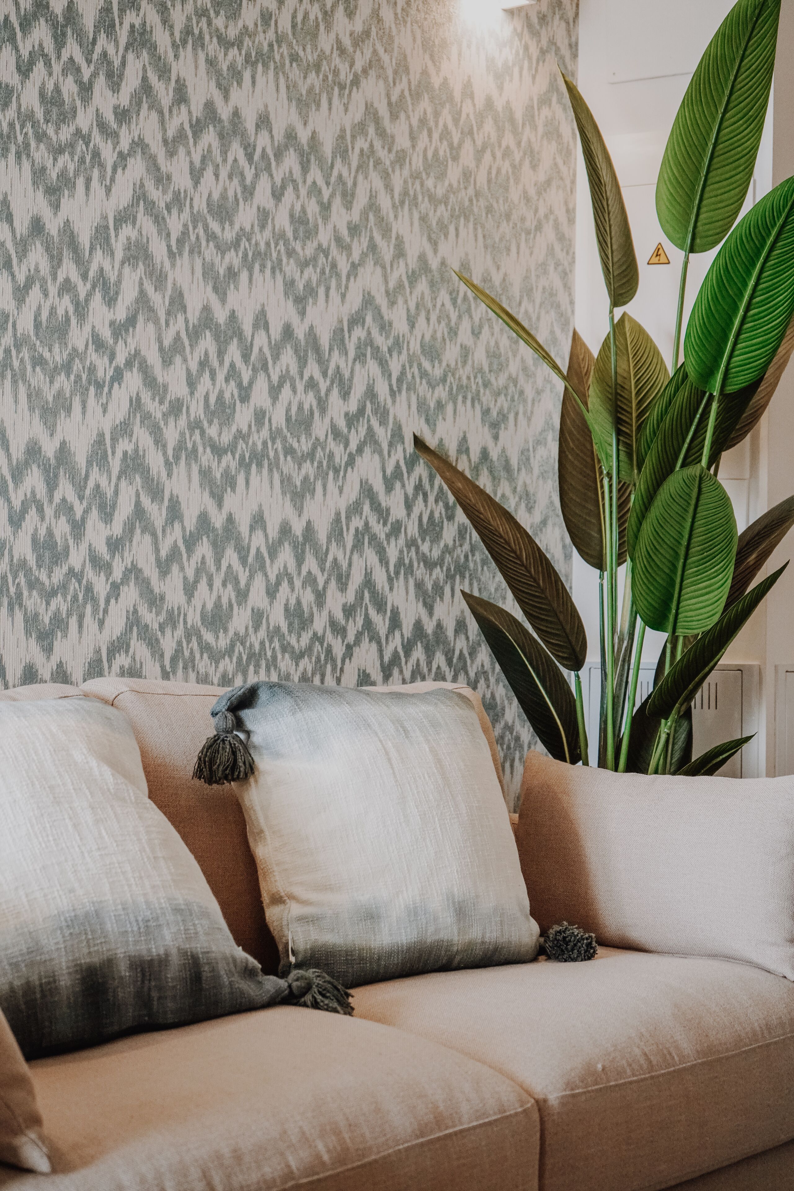 Un sofá beige con dos almohadas, una planta verde grande y un papel tapiz estampado.