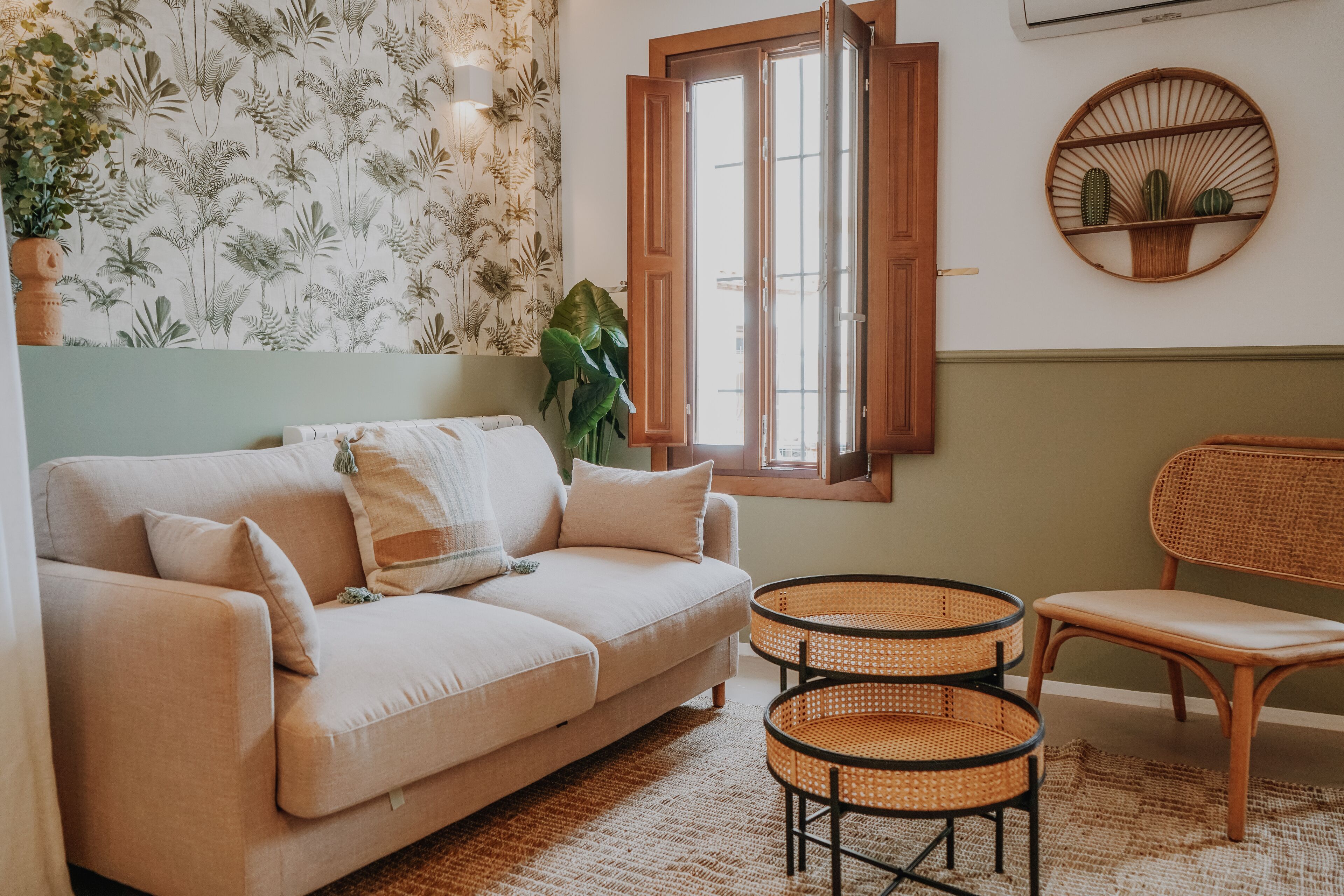 A living room with a beige sofa, two wicker chairs, a large plant, a wall-mounted cactus display, and a window with wooden shutters.