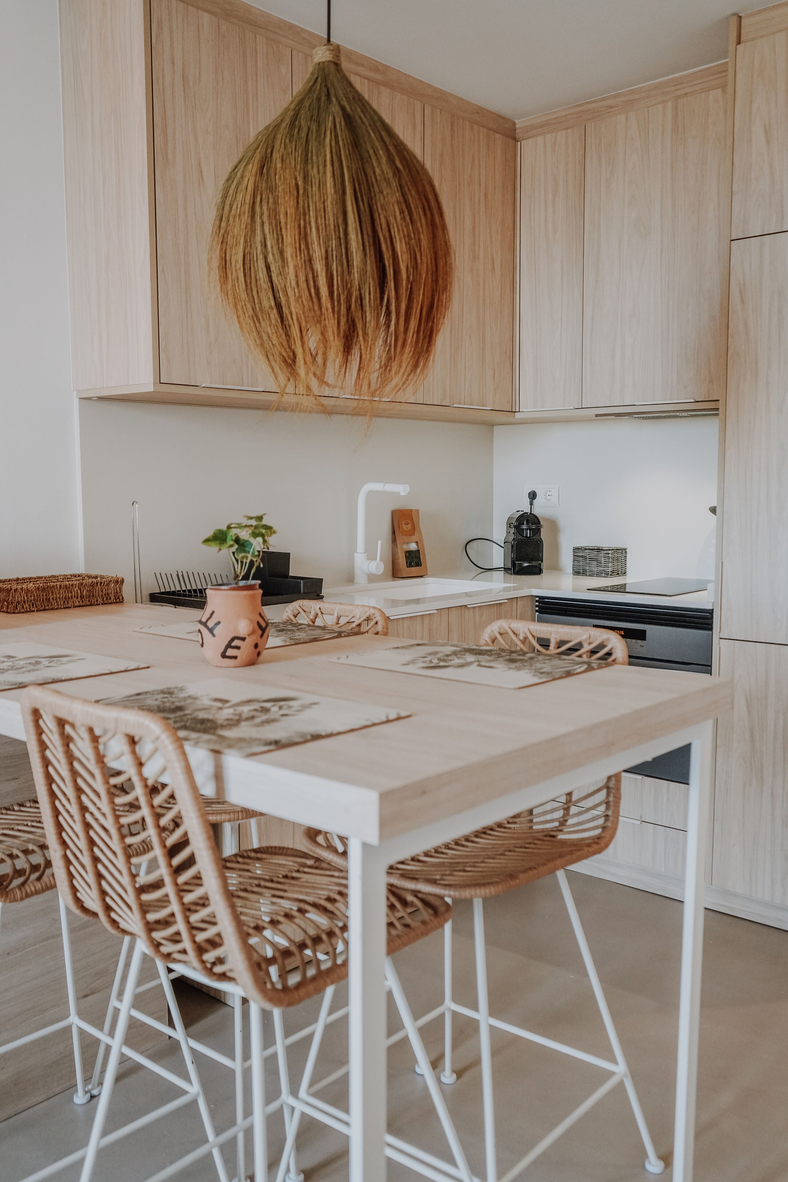 Una cocina moderna con armarios de madera clara, una lámpara colgante, una mesa de comedor con sillas y una cafetera.
