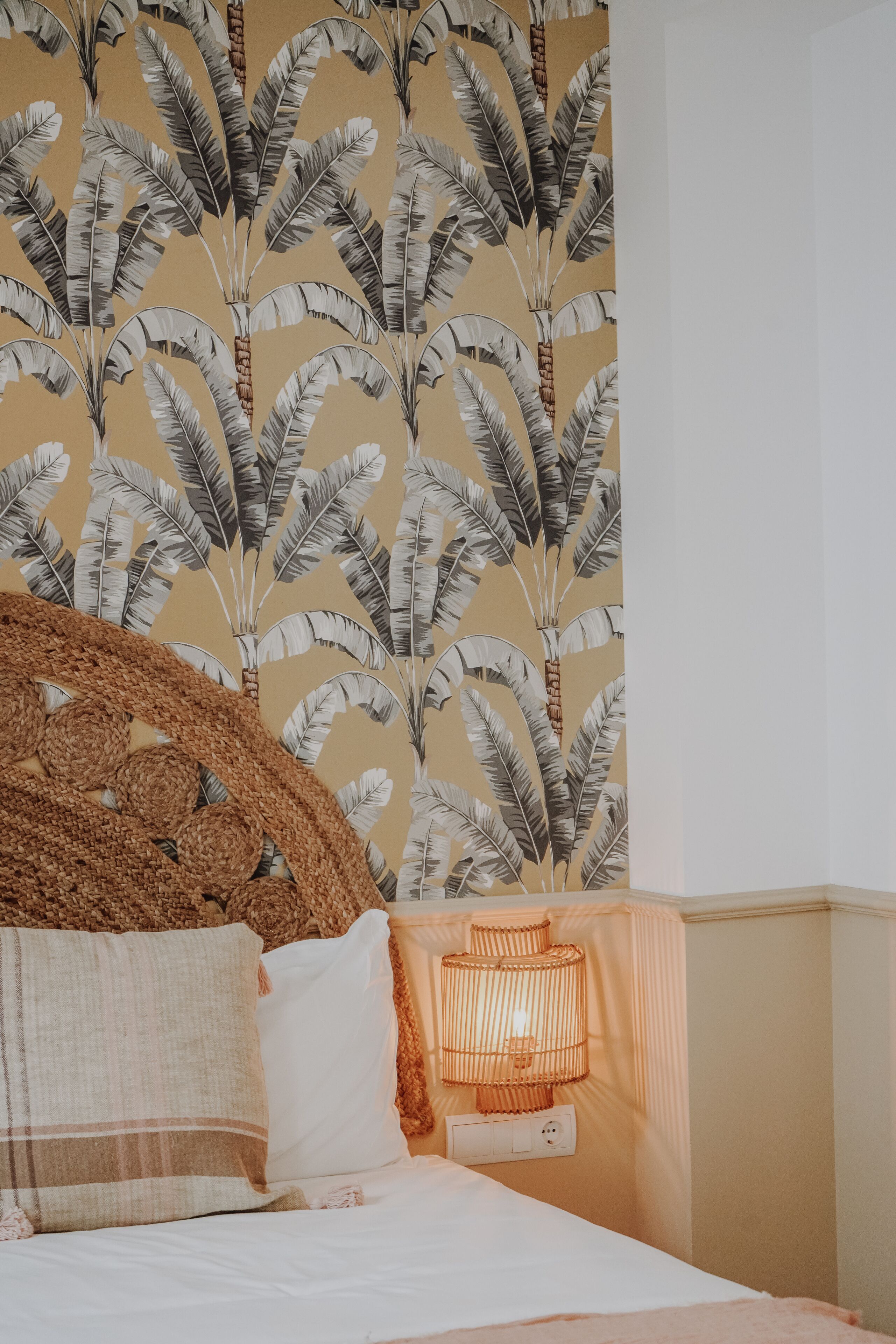 A bedroom with a bed, pillows, a woven headboard, a wall with a banana leaf pattern, and a bedside lamp.