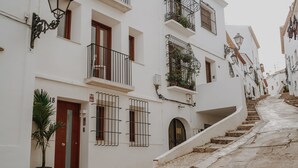 Una calle estrecha con edificios blancos, balcones con barandillas metálicas y una planta en maceta cerca de la entrada.