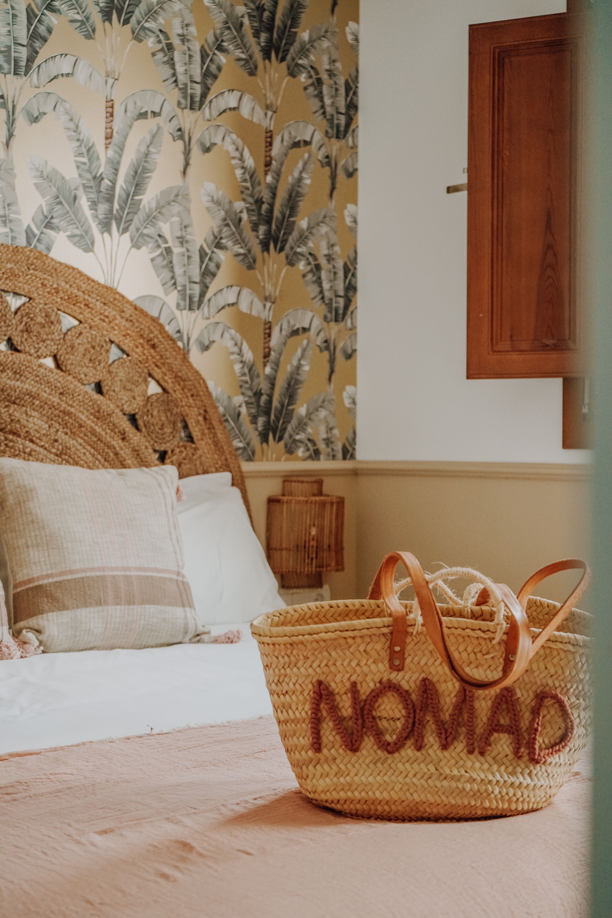 Un lit avec une tête de lit en osier, un panier en osier portant l ’inscription « NOMAD », et un mur orné d’un motif de feuilles tropicales.
