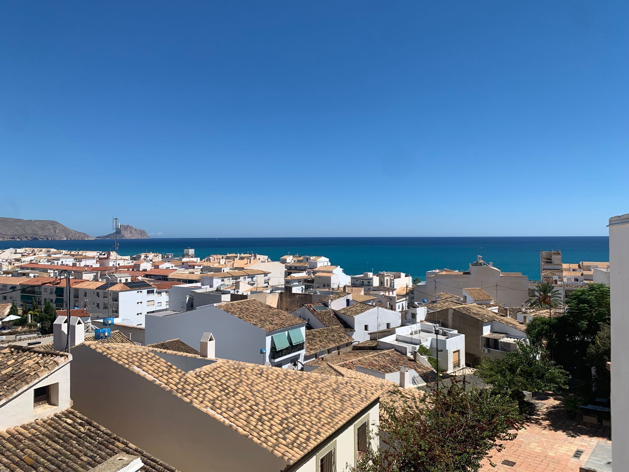 A coastal town with white buildings and terracotta roofs, a clear blue sky, and the sea in the background.
