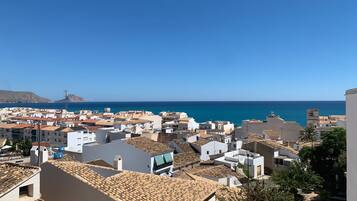 A coastal town with white buildings and terracotta roofs, a clear blue sky, and the sea in the background.