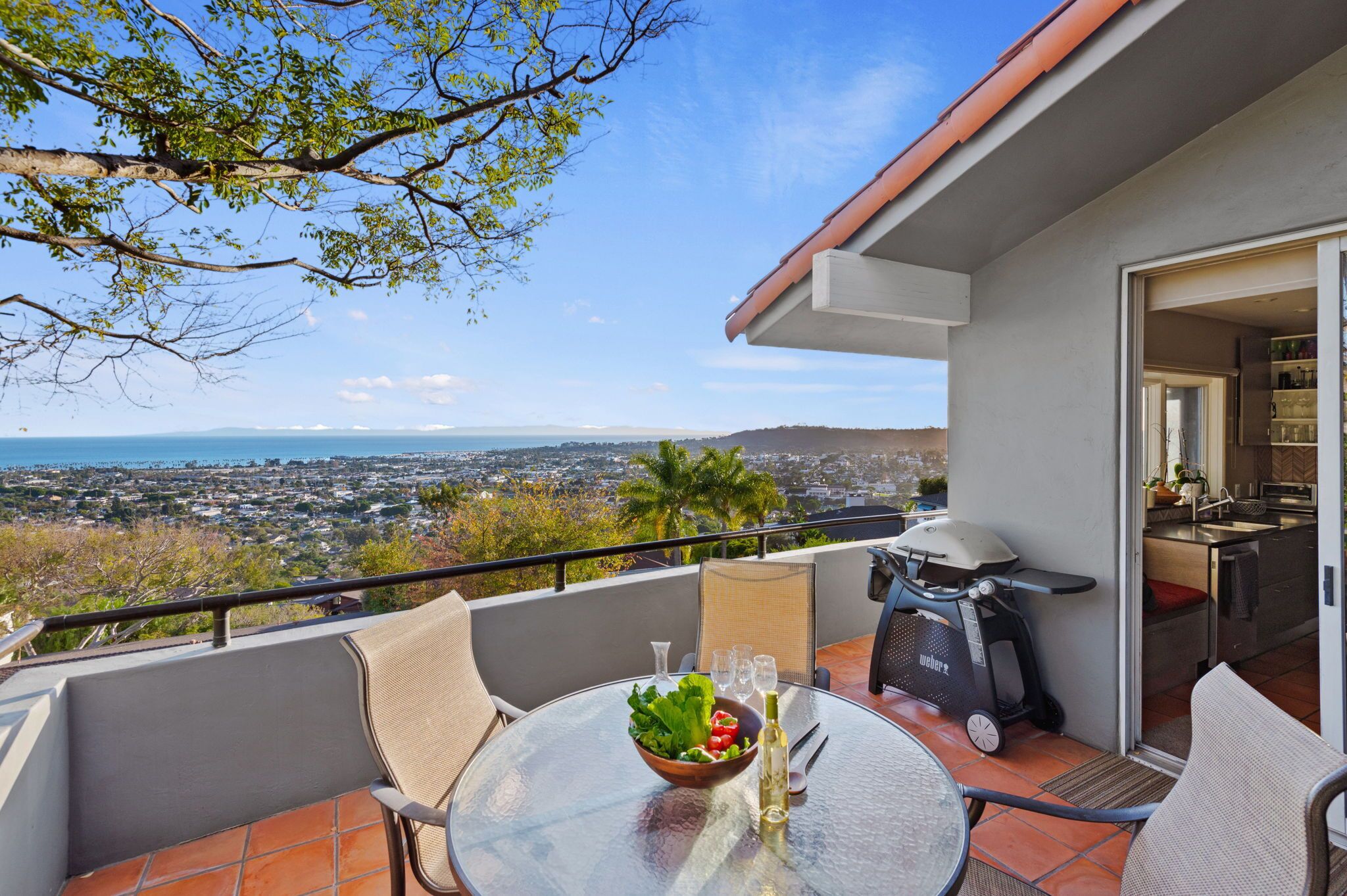 A balcony with a glass table set for dining, a grill, and a view of a cityscape.