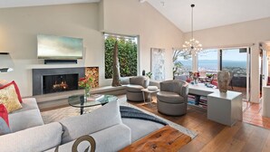 A spacious living room with a fireplace, comfortable seating, a coffee table, and a large window offering a view of the city. - Stunning Panoramic Views From Contemporary Riviera Home! (Santa Barbara)