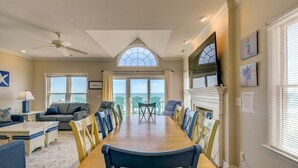 Un'ampia sala da pranzo con un lungo tavolo di legno e sedie, una ventola a soffitto e ampie finestre che offrono vista sull'oceano.