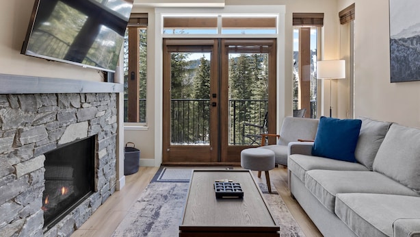 Un salón con chimenea de piedra, televisor de pantalla plana, sofá gris y balcón con vistas a árboles.