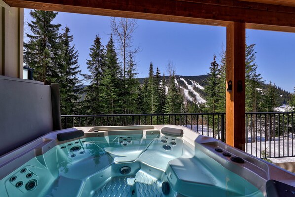 A hot tub with a wooden deck and a view of a snowy mountain and trees.