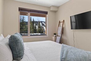 A bedroom with a large bed, a window with a view of trees and mountains, a flat-screen TV, and a painting on the wall.