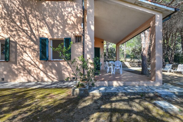 A covered outdoor area with stone flooring, a small tree, and white plastic chairs.