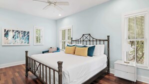 Une chambre à coucher comprenant un lit, une table de chevet, un ventilateur de plafond et deux fenêtres équipées de stores.