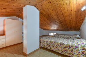 A bedroom with a sloped wooden ceiling, a bed with a polka dot bedspread, a white wardrobe, and a small bedside table with a lamp.