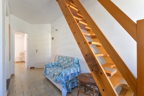 A room with a wooden staircase, a bed with a nautical-themed bedspread, a wicker chair, and a door leading to another room.