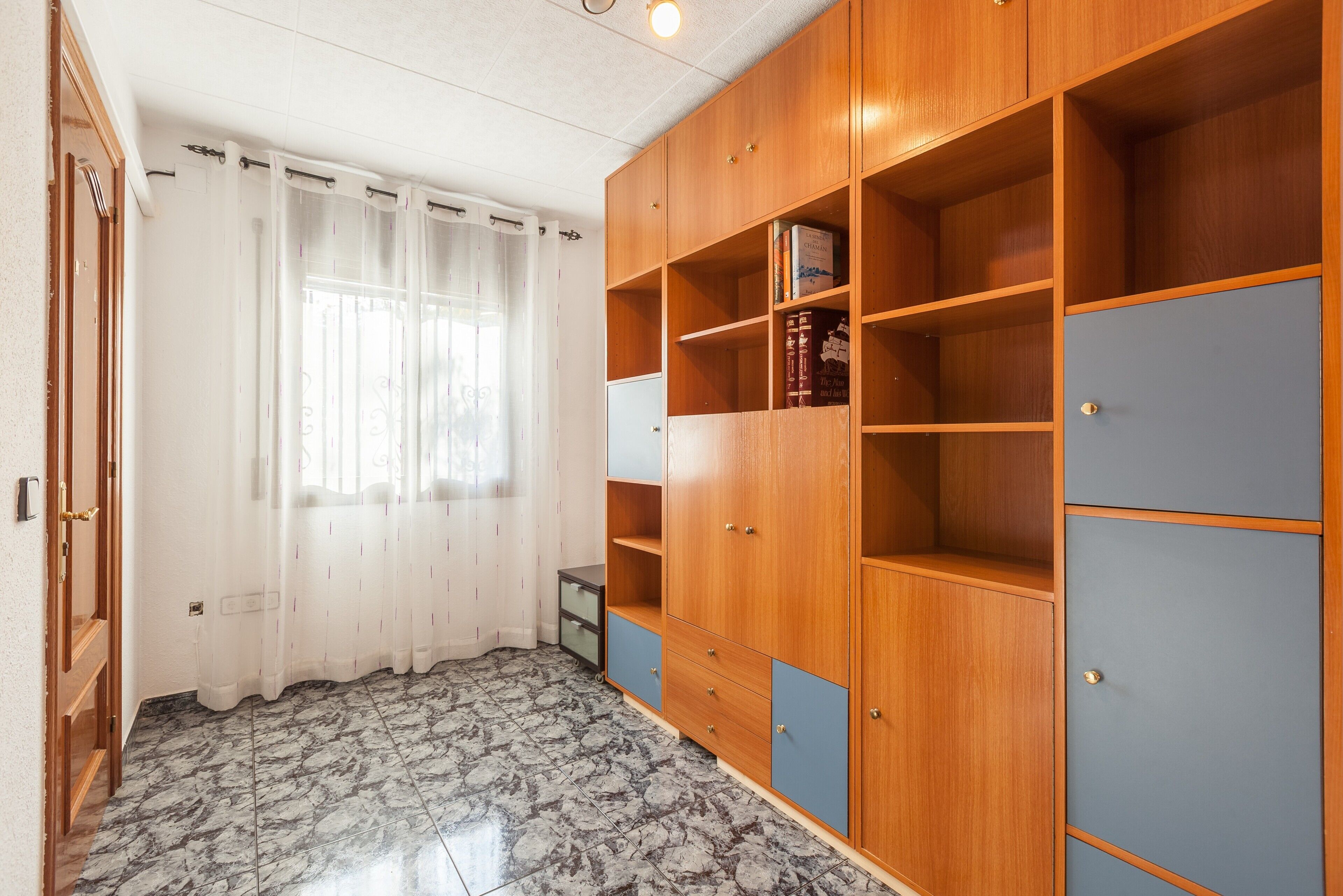 Quarto com guarda-roupa de madeira, janela com cortinas brancas e chão ladrilhado.