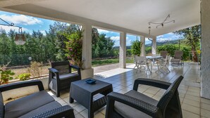 An outdoor patio area with wicker furniture, a table, and chairs, surrounded by greenery and a view of the landscape.