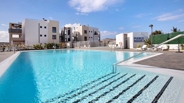 Ein Swimmingpool mit klarem, blauem Wasser, ein modernes Gebäude mit der Beschilderung 'PUEBLO MAJOR' und ein grüner Zaun im Hintergrund.