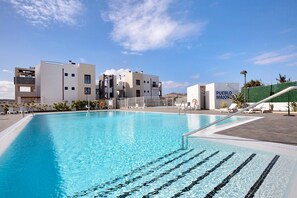 A swimming pool with clear blue water, a modern building with 'PUEBLO MAJOR' signage, and a green fence in the background.