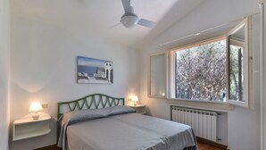 Un dormitorio con cama, dos lámparas de noche, ventilador de techo, ventana con vista a árboles y un cuadro en la pared.