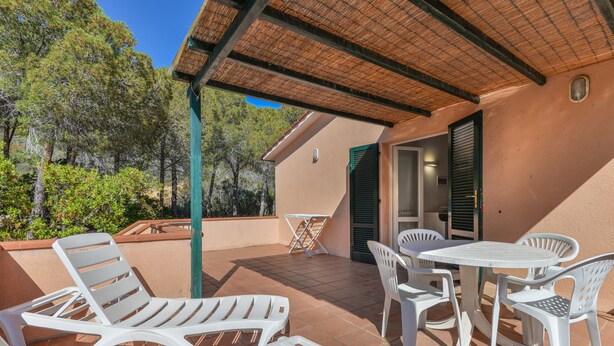Un patio exterior con sillas de plástico blancas y una mesa, rodeado por una pérgola y un edificio con tejado de baldosas.