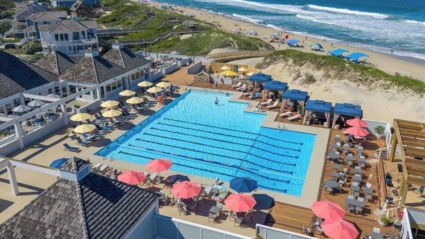 A swimming pool with sun loungers and umbrellas, a beach with people, and a view of the ocean. - Sunset Retreat 2 Bedroom Pet-Friendly Condo at Oyster Point (Kill Devil Hills)