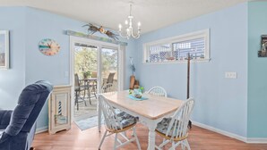 A dining area with a wooden table and white chairs, a chandelier, and a view of the outdoors through a glass door. - Sunset Retreat 2 Bedroom Pet-Friendly Condo at Oyster Point (Kill Devil Hills)