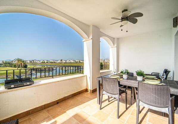 A balcony with a view of a golf course and water, a table with chairs, and a ceiling fan.