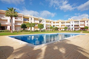 A swimming pool surrounded by a courtyard with multi-story buildings, a palm tree, and a clear blue sky. - Apartment "Columbus Beach" with Sea View, Shared Pool & Wi-Fi (Quarteira)