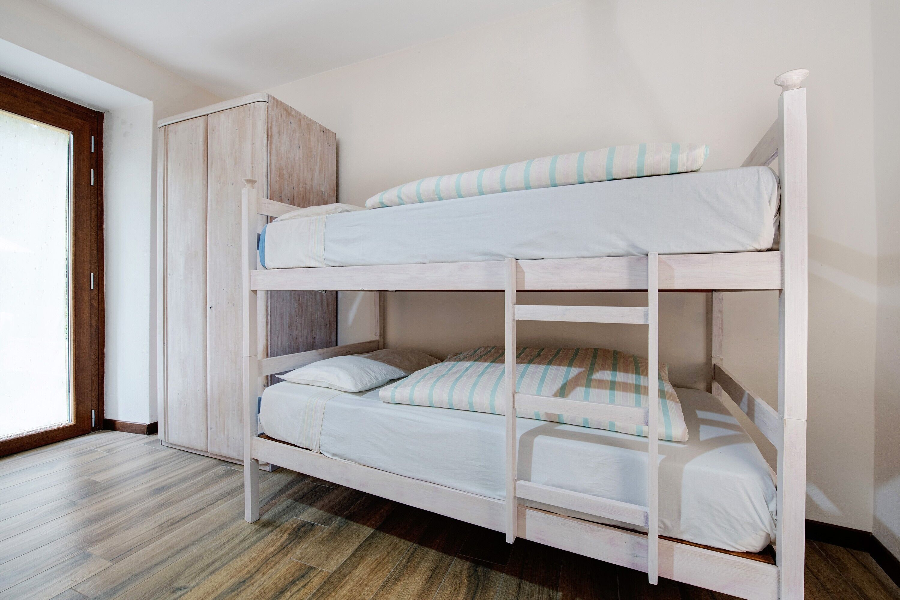 Una cama litera con un armario de madera, una ventana con persianas y suelo de madera.