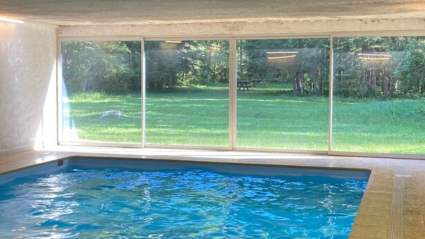 Une piscine intérieure  à l’eau claire, avec vue sur une cour verdoyante grâce à de grandes fenêtres, et un sol carrelé.