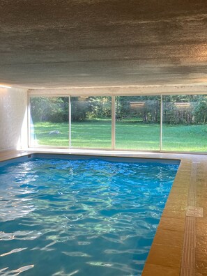 Une piscine intérieure à l’eau claire, avec vue sur une cour verdoyante grâce à de grandes fenêtres, et un sol carrelé.