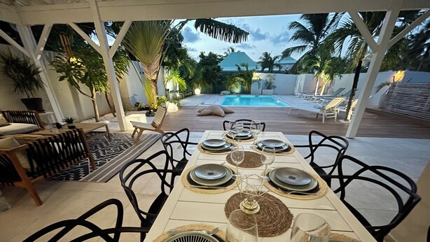 Un área de comedor al aire libre con vista a la piscina, mesas dispuestas con platos y vasos, y plantas tropicales.