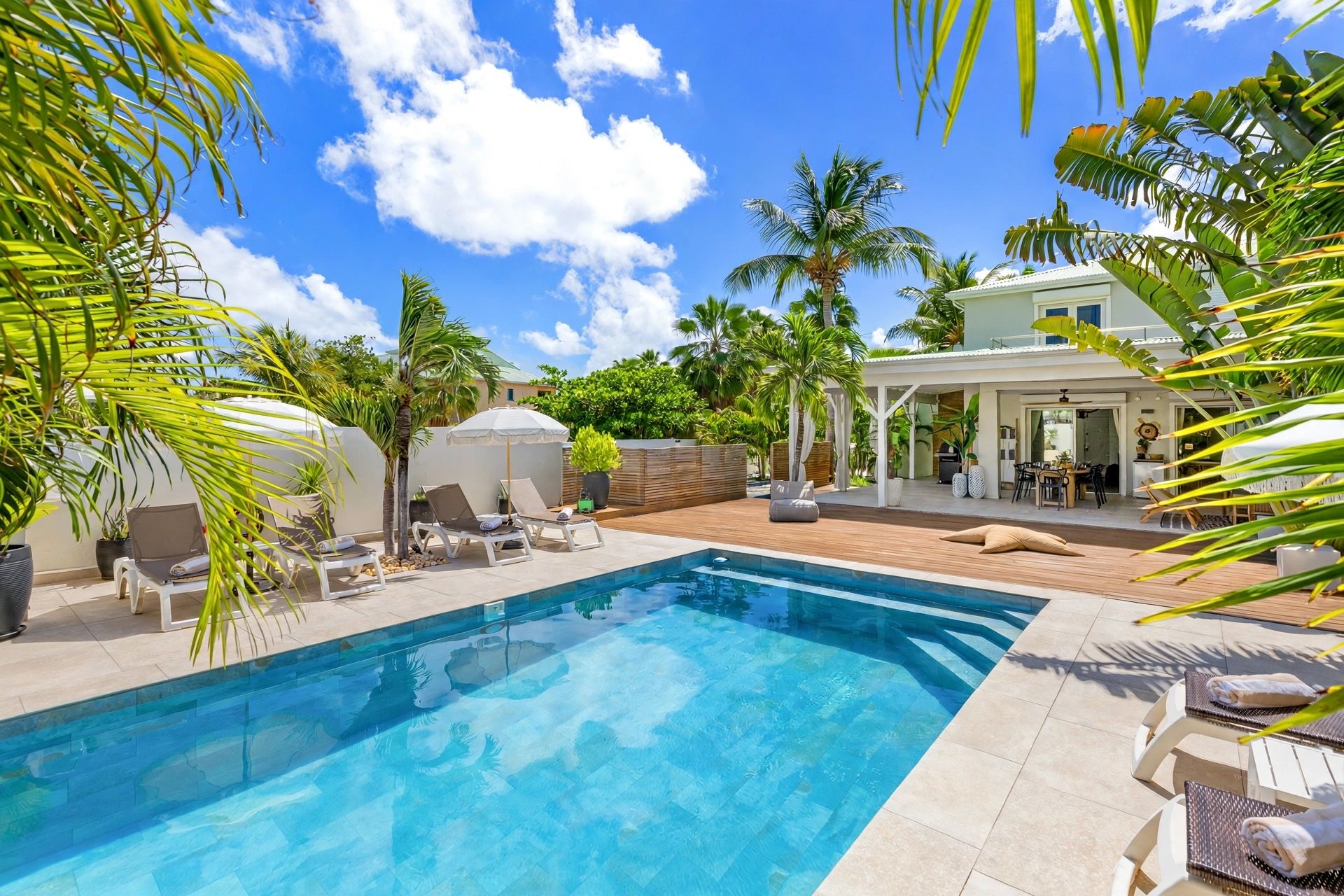 A swimming pool with lounge chairs and umbrellas, surrounded by palm trees and a patio area.